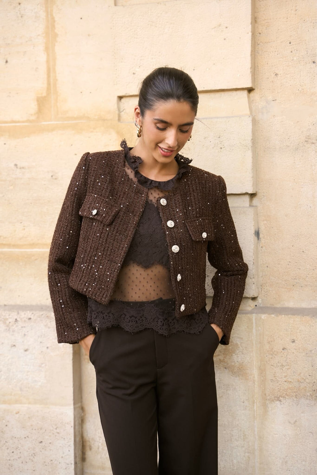 A woman wearing a cropped brown tweed jacket with silver-tone buttons and sequins, layered over a sheer black polka-dot top and black pants, standing against a light stone wall.