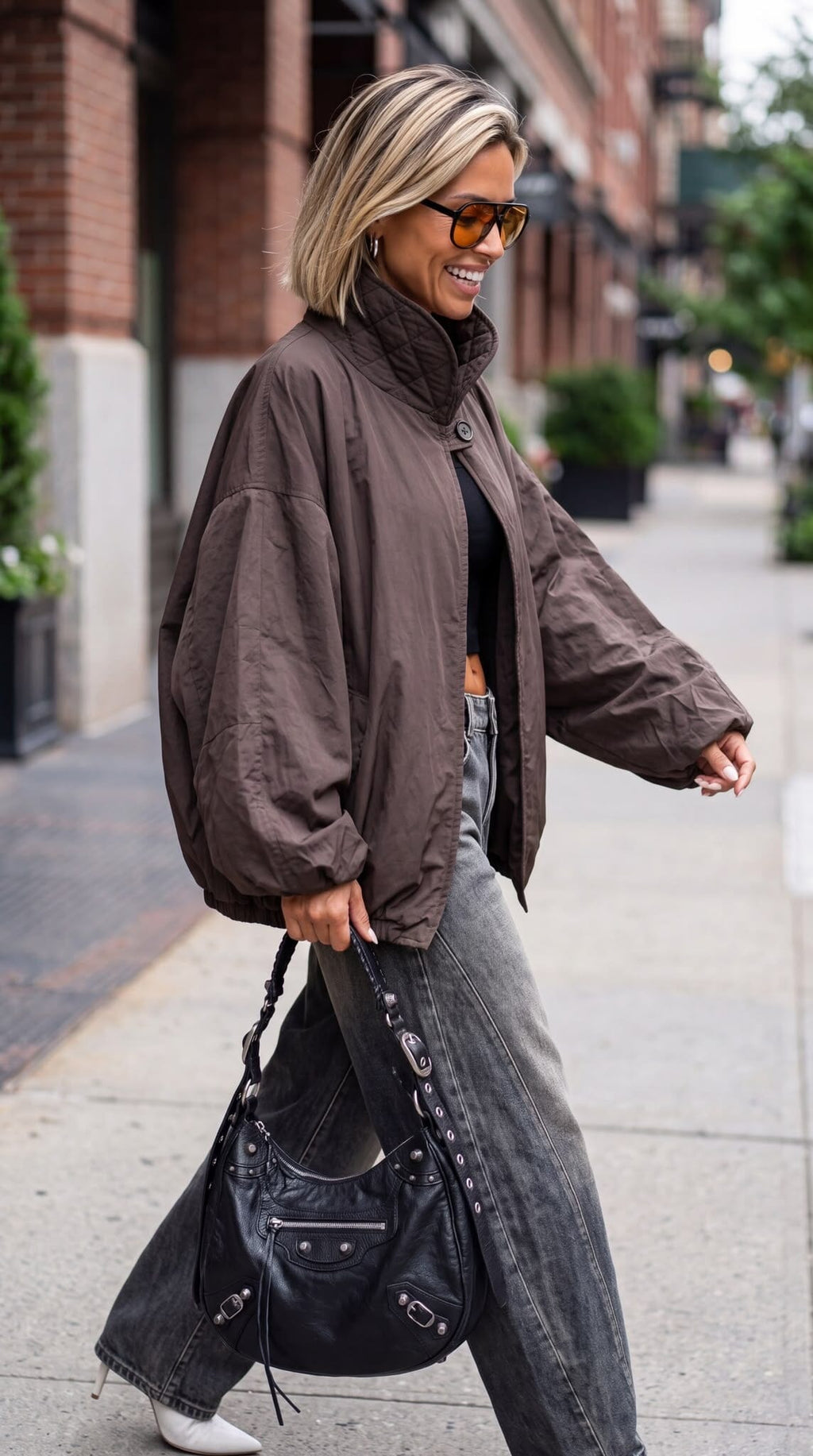 A woman models an oversized dark brown bomber jacket, styled with dark gray baggy jeans, a yellow top, white sneakers, and a black leather shoulder bag against a textured beige wall.